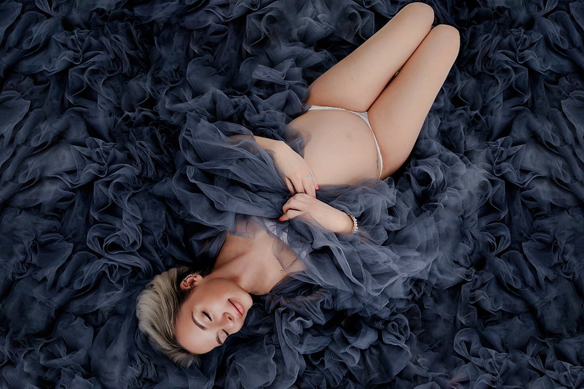 Femme enceinte allongée dans un voilage bleu nuit lors d’une séance photo grossesse artistique en studio.