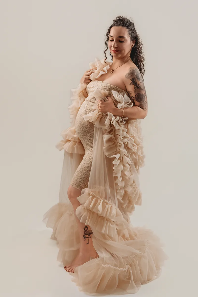 Future maman en robe de dentelle beige et voilage à volants lors d’une séance photo grossesse en studio.