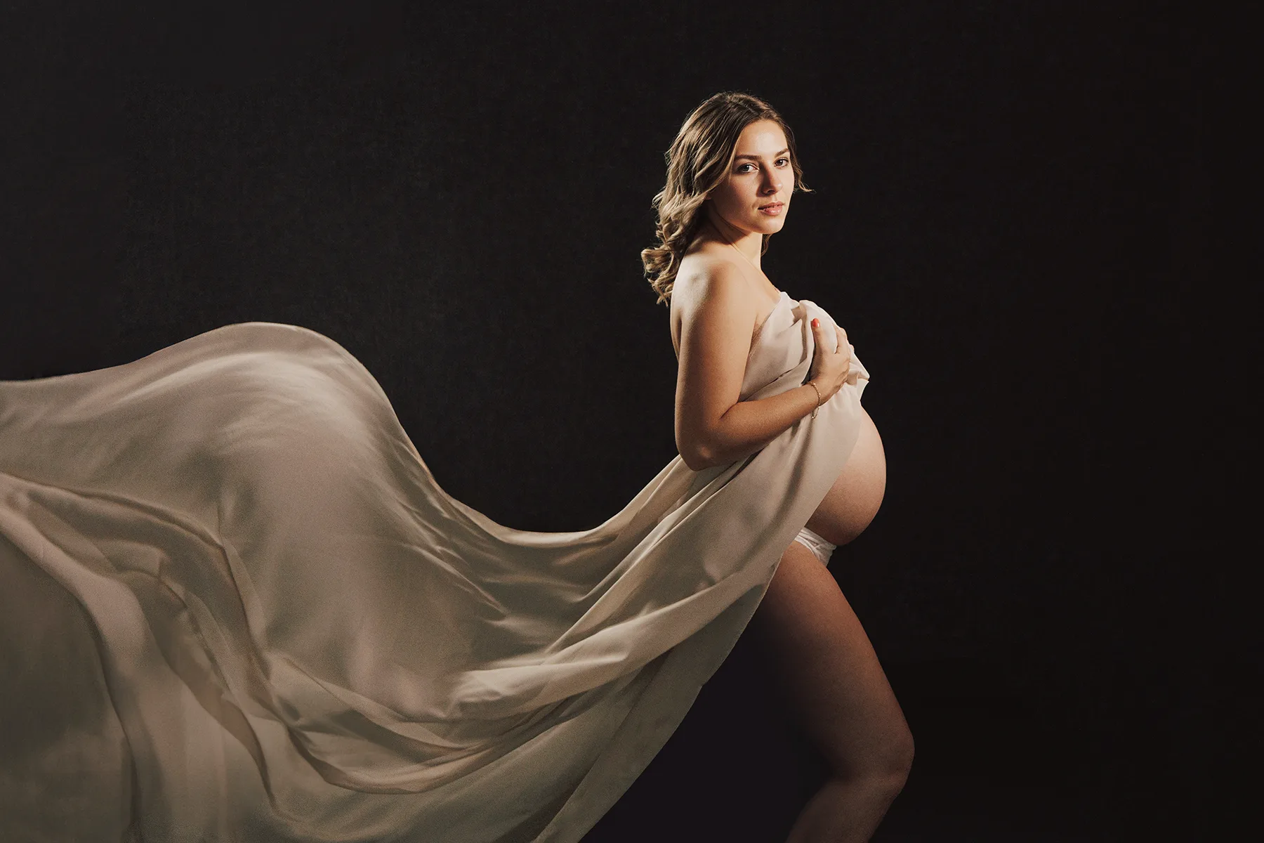 Femme enceinte drapée d’un tissu beige lors d’une séance photo grossesse artistique en studio.