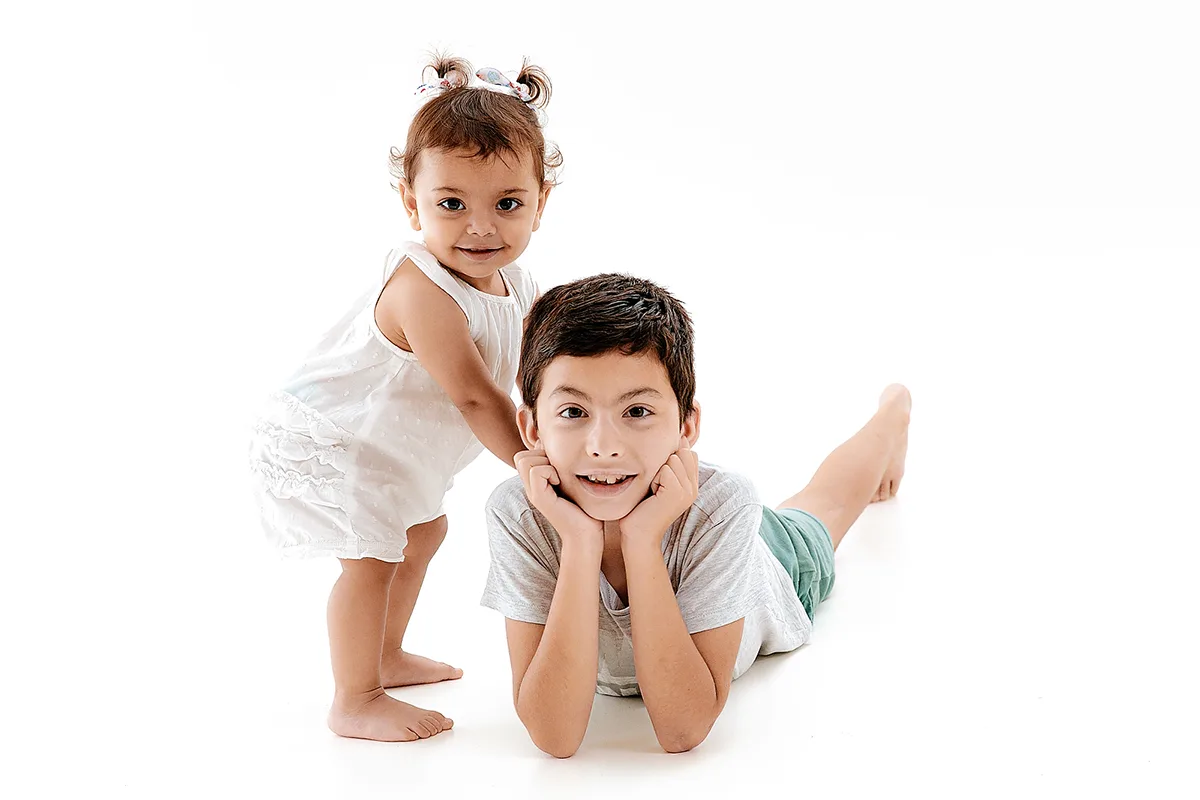 Frère et petite sœur souriants photographiés sur fond blanc lors d’une séance photo en studio.