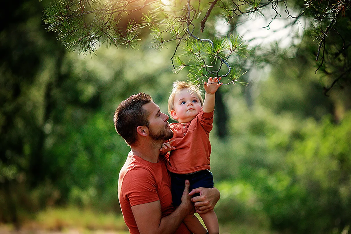 Papa portant son jeune enfant qui touche une branche lors d’une séance photo en famille dans la nature.