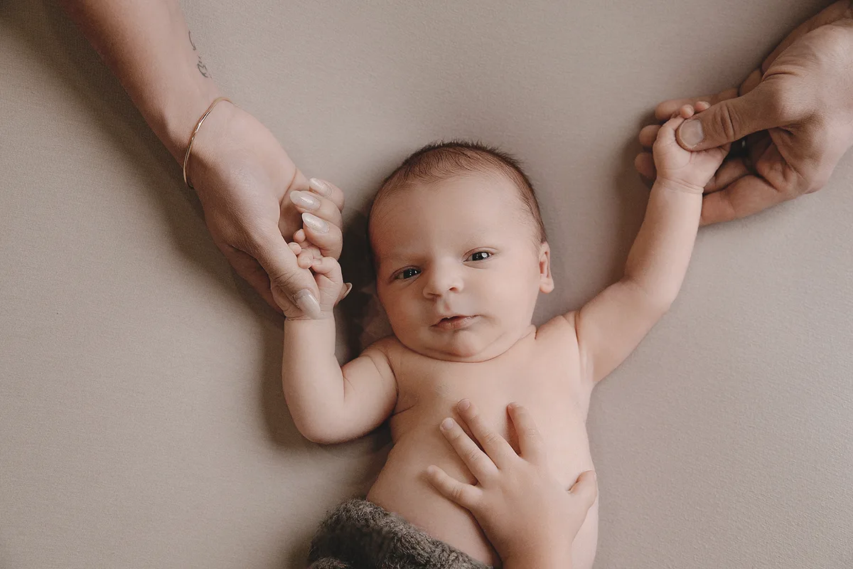 Nouveau-né éveillé tenant les doigts de ses parents lors d’une séance photo naissance en studio.