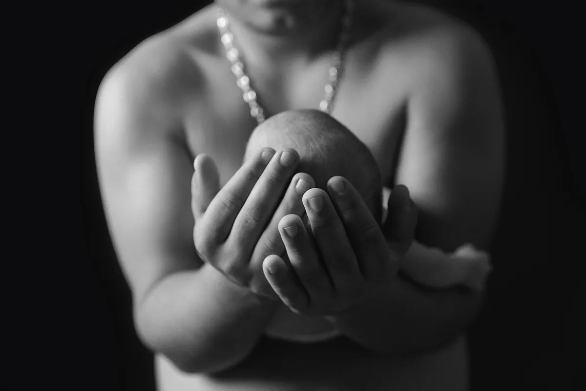 Papa tenant délicatement son nouveau-né dans ses mains lors d’un portrait artistique en noir et blanc.