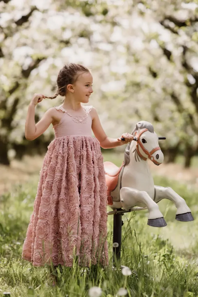 Jeune fille en robe rose près d’un cheval à bascule, photographiée sous les arbres en fleurs en Provence.